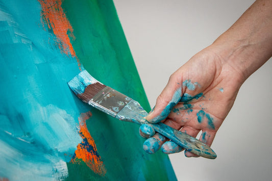 Artists hand on paintbrush adding teal paint to the start of an abstract painting