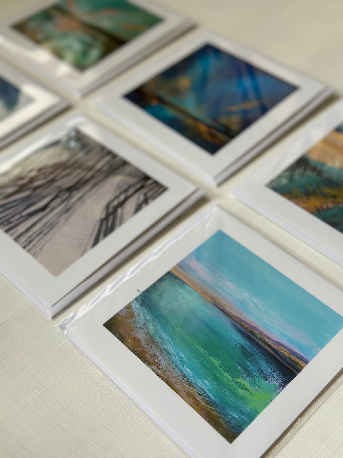 A set of art print cards arranged on a light surface, showing abstract landscape and seascape paintings with white borders and varying color palettes.