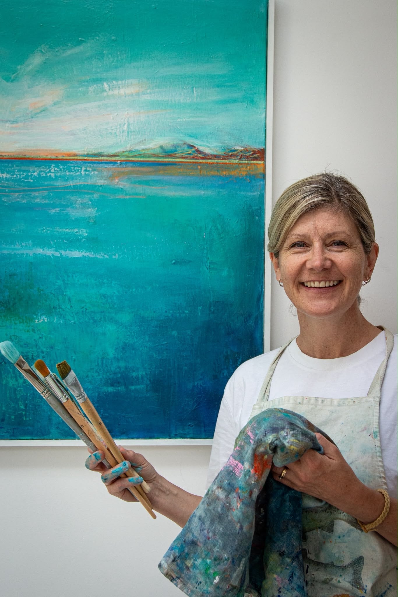 Helen Wooldridge artist wearing a white t-shirt and paint-splattered apron holds several paintbrushes and a canvas while standing in front of a large blue and teal landscape painting on a white wall.