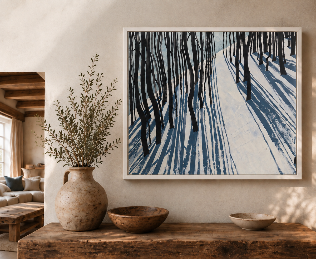 A framed fine art canvas print ‘Snow Shadow’ by Helen Wooldridge depicting a snowy forest with dark tree trunks and blue snow shadows, displayed on a beige wall above a rustic wooden console table with a ceramic vase and bowls.