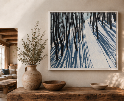 A framed fine art canvas print ‘Snow Shadow’ by Helen Wooldridge depicting a snowy forest with dark tree trunks and blue snow shadows, displayed on a beige wall above a rustic wooden console table with a ceramic vase and bowls.