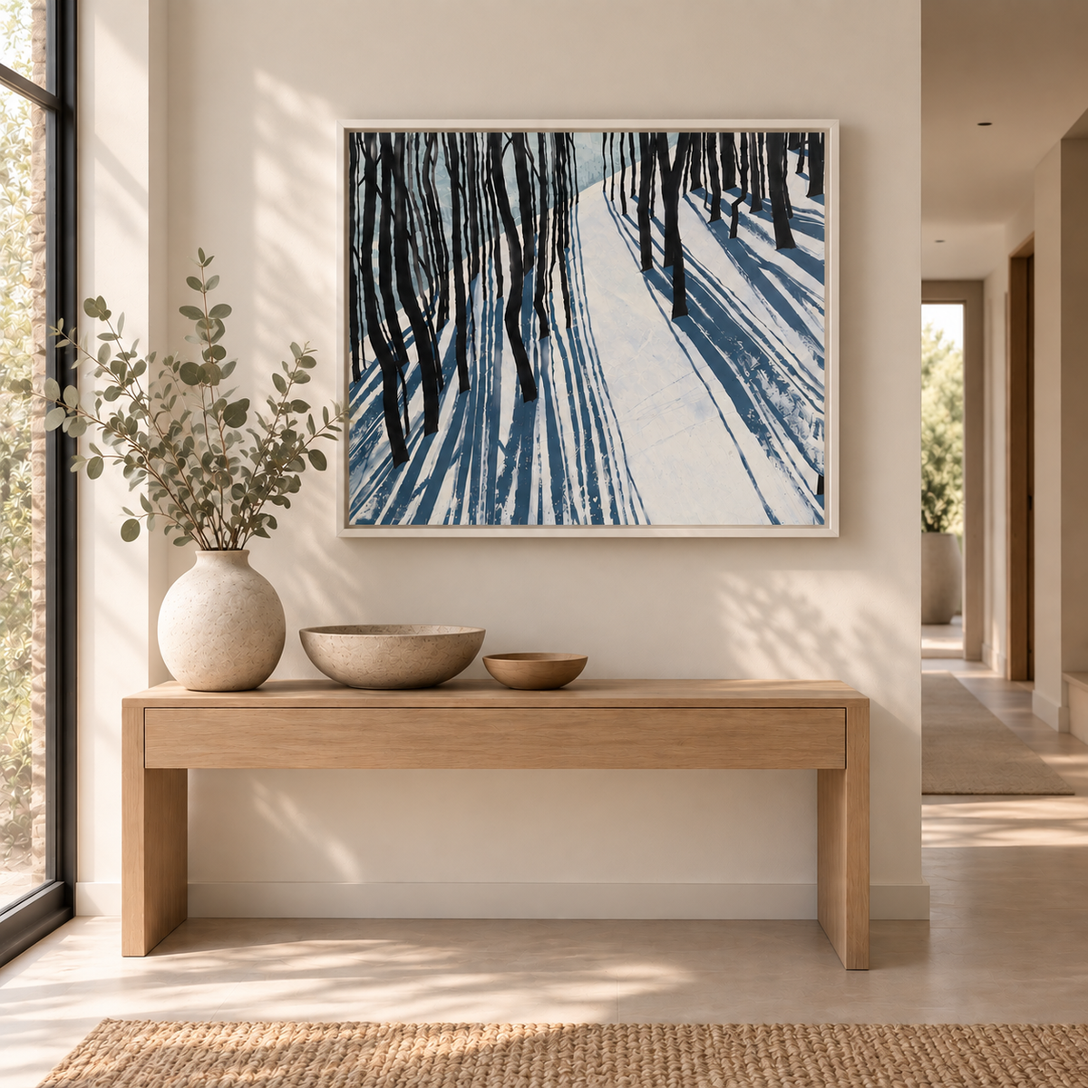 A framed fine art canvas print of a snow woodland painting by Helen Wooldridge with light on snow through trees. Hung in a contemporary interior with console table in light hallway.