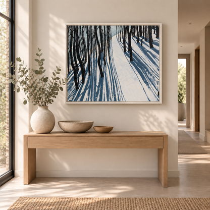 A framed fine art canvas print of a snow woodland painting by Helen Wooldridge with light on snow through trees. Hung in a contemporary interior with console table in light hallway.