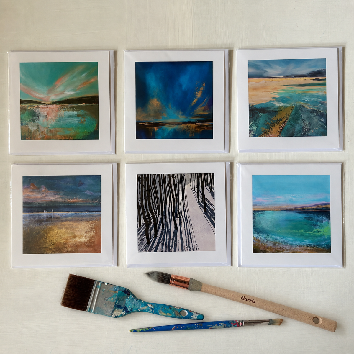 Six landscape art prints presented as greeting cards arranged in a 2x3 grid on a light beige surface, with three paintbrushes placed below. The paintings depict colorful water scenes, a snowy forest path, and dramatic sky landscapes in blue, teal, green, gold, and brown tones.