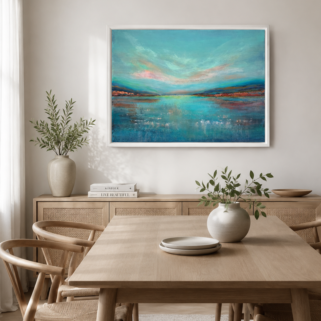 A framed abstract landscape painting with turquoise, blue, and pink tones hangs on a light beige wall above a wooden dining table and chairs in a styled interior room.
